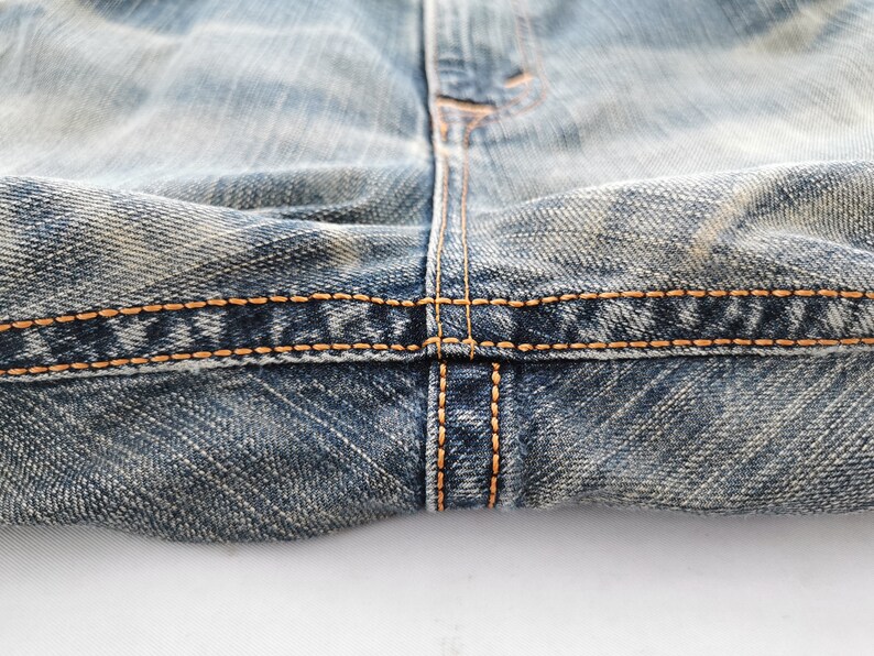 Puede incluir: Primer plano de unos vaqueros de mezclilla azul con costuras en hilo naranja-amarillo. La imagen destaca los detalles de las costuras y la textura de la tela vaquera desgastada.