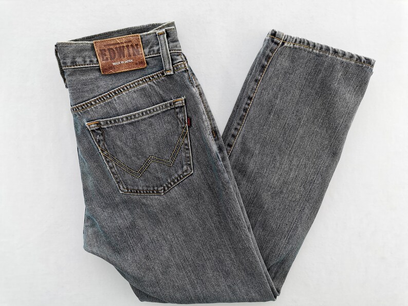May include: Gray denim jeans with a classic five-pocket design. Features a brown leather patch with the brand name and a red tag on the back pocket. The jeans are made of a durable fabric and have a straight leg cut.