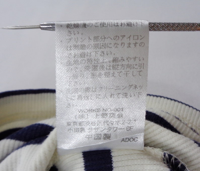 May include: A white fabric garment care label with Japanese text printed in black, featuring instructions for washing and maintenance. The label is attached to a ribbed knit textile with alternating horizontal navy blue and white stripes.