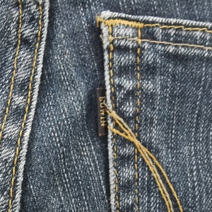 K&ouml;nnte beinhalten: Dunkelblauer Denim-Stoff mit einer Ges&auml;&szlig;tasche, die mit dicken, goldgelben Kontrastn&auml;hten versehen ist. Ein kleines, rechteckiges, dunkelbraunes Stoffetikett ist in die Seitennaht der Tasche eingen&auml;ht und zeigt das Wort EDWIN in goldenen Gro&szlig;buchstaben.