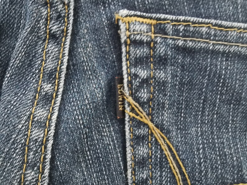 K&ouml;nnte beinhalten: Dunkelblauer Denim-Stoff mit einer Ges&auml;&szlig;tasche, die mit dicken, goldgelben Kontrastn&auml;hten versehen ist. Ein kleines, rechteckiges, dunkelbraunes Stoffetikett ist in die Seitennaht der Tasche eingen&auml;ht und zeigt das Wort EDWIN in goldenen Gro&szlig;buchstaben.