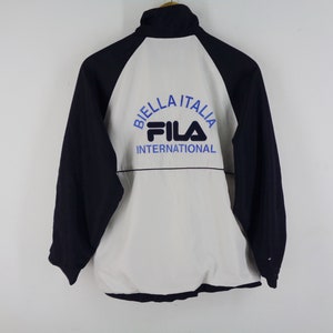 May include: A navy blue, white, and light blue Fila International jacket. The jacket has a white front and back panel with navy blue sleeves. The text "BIELLA ITALIA" and "FILA INTERNATIONAL" are printed on the back in blue.
