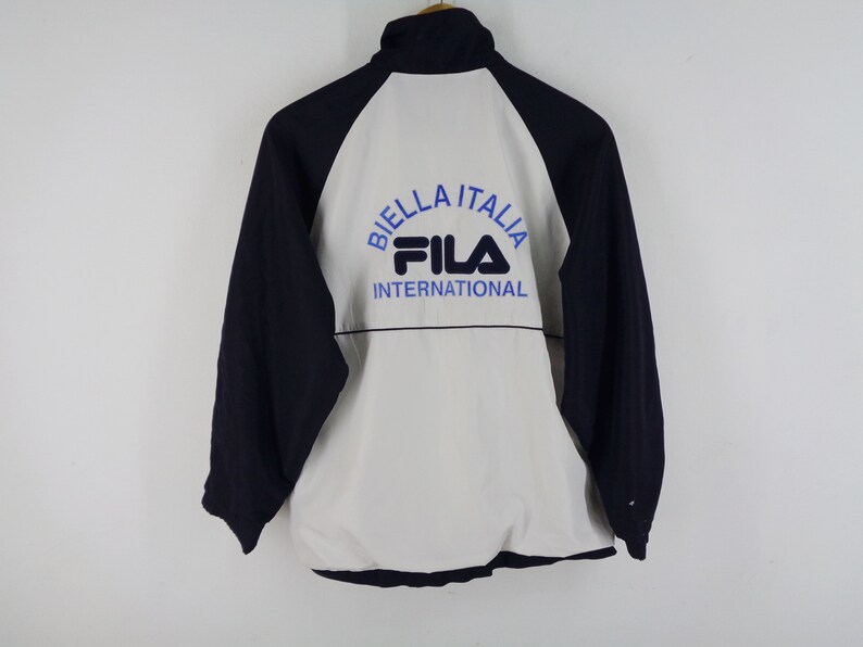 May include: A navy blue, white, and light blue Fila International jacket. The jacket has a white front and back panel with navy blue sleeves. The text "BIELLA ITALIA" and "FILA INTERNATIONAL" are printed on the back in blue.
