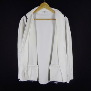 May include: A white zip-up jacket with navy blue trim and two front pockets. The jacket is hanging on a wooden hanger against a black background. The jacket has a textured fabric.