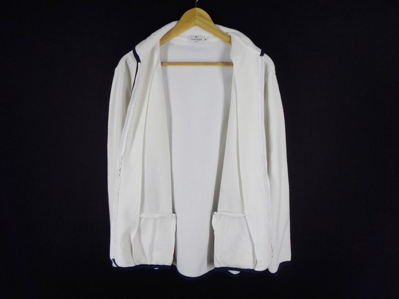May include: A white zip-up jacket with navy blue trim and two front pockets. The jacket is hanging on a wooden hanger against a black background. The jacket has a textured fabric.