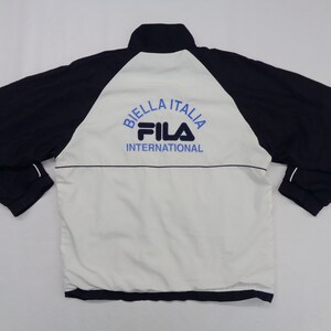 May include: A navy blue and white Fila jacket with the text "BIELLA ITALIA" and "FILA INTERNATIONAL" in blue. The jacket has a high collar and long sleeves.