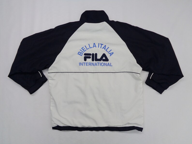 May include: A navy blue and white Fila jacket with the text "BIELLA ITALIA" and "FILA INTERNATIONAL" in blue. The jacket has a high collar and long sleeves.