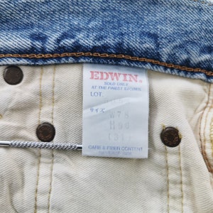 May include: Close-up of the inside of a pair of blue denim jeans. The jeans have a white interior and a tag with the brand name "EDWIN" and size information. The jeans have brown metal buttons.