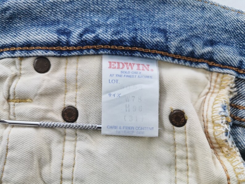 May include: Close-up of the inside of a pair of blue denim jeans. The jeans have a white interior and a tag with the brand name "EDWIN" and size information. The jeans have brown metal buttons.