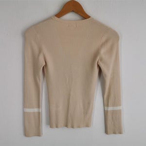 May include: A beige ribbed knit sweater with long sleeves, hanging on a wooden hanger. The sleeves have a white stripe detail near the cuffs. The sweater has a round neckline.
