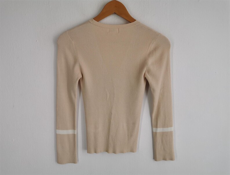 May include: A beige ribbed knit sweater with long sleeves, hanging on a wooden hanger. The sleeves have a white stripe detail near the cuffs. The sweater has a round neckline.