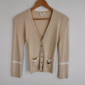 May include: A beige ribbed knit cardigan with a V-neck, long sleeves, and silver buttons. The cardigan has two front pockets with white trim and a decorative chain detail on one pocket. The sleeves have white stripes near the cuffs.