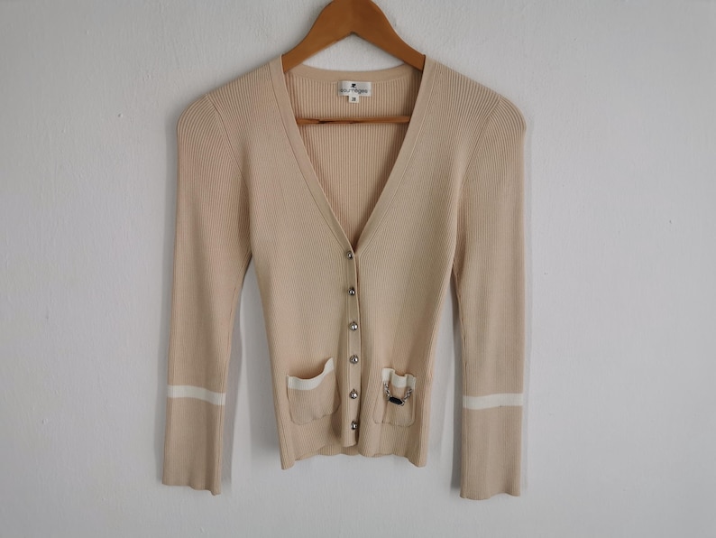 May include: A beige ribbed knit cardigan with a V-neck, long sleeves, and silver buttons. The cardigan has two front pockets with white trim and a decorative chain detail on one pocket. The sleeves have white stripes near the cuffs.