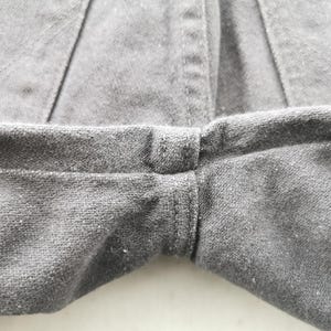 May include: Close-up of gray fabric, likely denim or a similar material, showing the seam where the legs of the garment meet. The stitching is visible, and the fabric has a slightly textured appearance. The overall tone is neutral and muted.