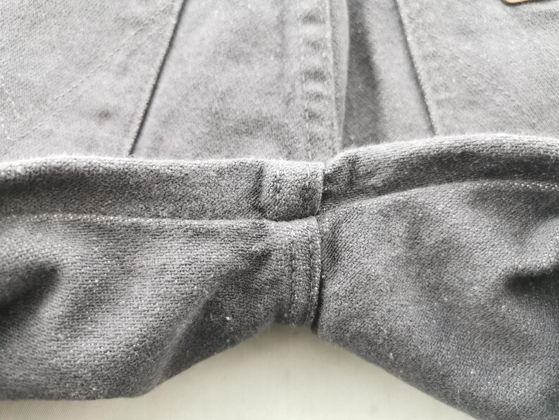 May include: Close-up of gray fabric, likely denim or a similar material, showing the seam where the legs of the garment meet. The stitching is visible, and the fabric has a slightly textured appearance. The overall tone is neutral and muted.
