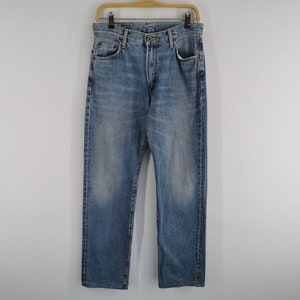 May include: Light blue denim jeans hanging on a wooden hanger. The jeans have a classic five-pocket design, a button and zipper closure, and a faded wash. The jeans are straight-legged and appear to be a medium weight.