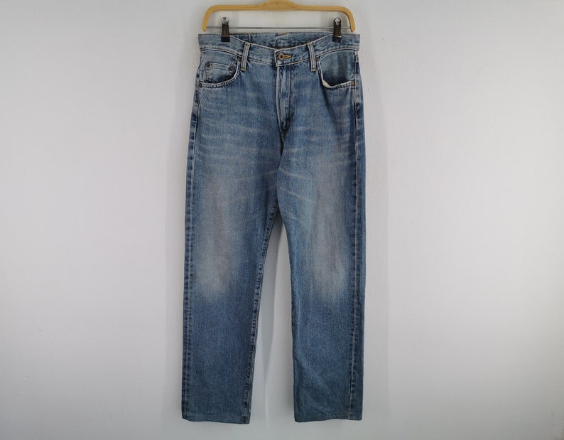 May include: Light blue denim jeans hanging on a wooden hanger. The jeans have a classic five-pocket design, a button and zipper closure, and a faded wash. The jeans are straight-legged and appear to be a medium weight.