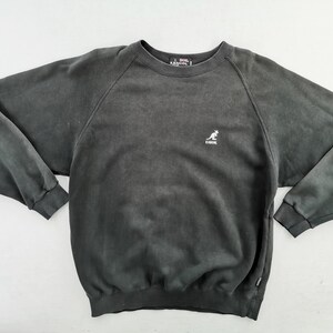 May include: A charcoal gray Kangol sweatshirt with a crew neck and long sleeves. The sweatshirt features a small white kangaroo logo on the chest. The fabric has a faded, vintage appearance.