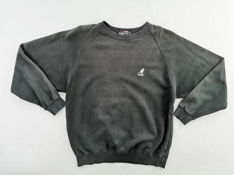 May include: A charcoal gray Kangol sweatshirt with a crew neck and long sleeves. The sweatshirt features a small white kangaroo logo on the chest. The fabric has a faded, vintage appearance.