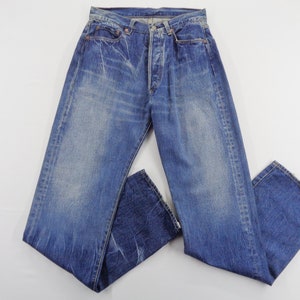 May include: Faded blue denim jeans with a button fly. The jeans have a classic five-pocket design and show signs of wear and fading, giving them a vintage look. The jeans are laid flat on a white surface.