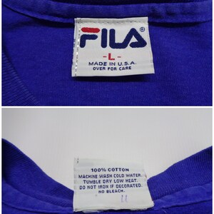 May include: Close-up of a blue FILA t-shirt label. The label reads "FILA" in red and navy blue, with "-L-" and "MADE IN U.S.A." below. The care instructions are also visible, including "100% COTTON" and washing instructions.