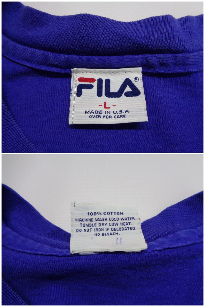 May include: Close-up of a blue FILA t-shirt label. The label reads "FILA" in red and navy blue, with "-L-" and "MADE IN U.S.A." below. The care instructions are also visible, including "100% COTTON" and washing instructions.
