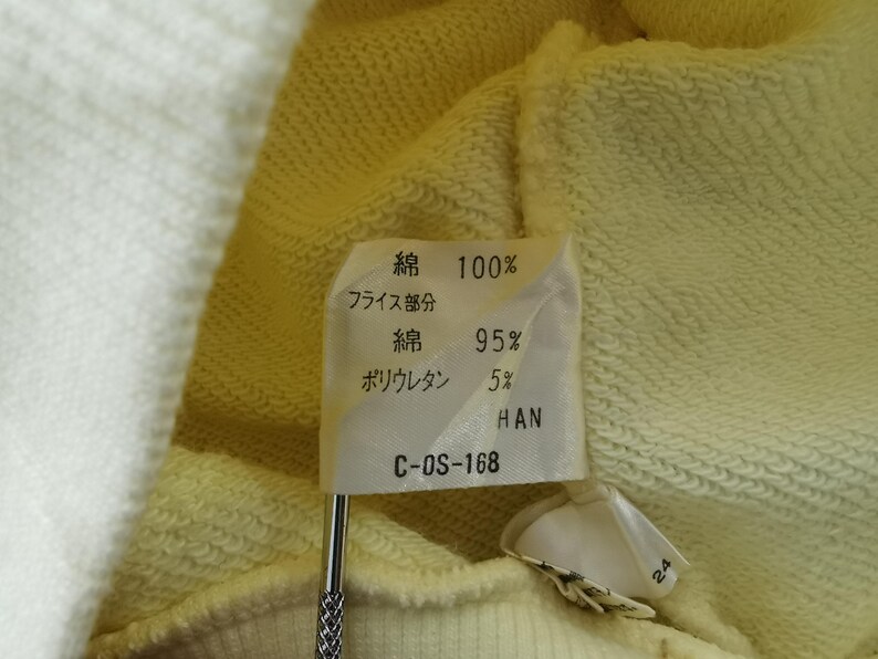 May include: Close-up of a fabric tag with Japanese text indicating the material composition: 100% cotton, 95% cotton, and 5% polyurethane. The tag is attached to a light yellow fabric. The tag also has the text "HAN" and "C-OS-168".