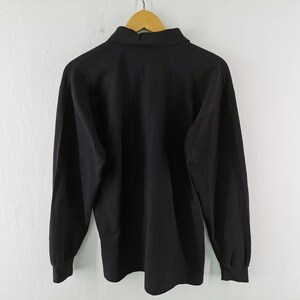 May include: A solid black long-sleeve shirt with a collared neckline, hanging on a wooden hanger. The shirt's fabric appears smooth, and the sleeves have cuffs. The background is a plain white wall.