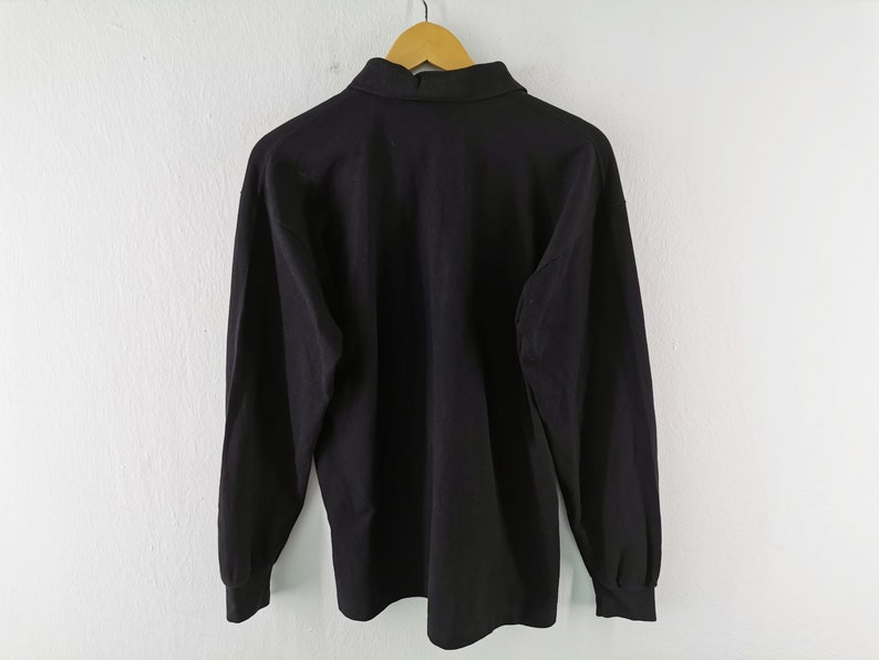May include: A solid black long-sleeve shirt with a collared neckline, hanging on a wooden hanger. The shirt's fabric appears smooth, and the sleeves have cuffs. The background is a plain white wall.