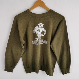 Puede incluir: Sudadera verde oliva con un gráfico blanco de una mascota musculosa con forma de búho que lleva una camiseta con la letra "A". El texto "AMERICAN UNIVERSITY" está impreso debajo. La sudadera tiene mangas largas y cuello redondo.