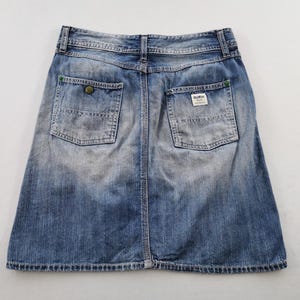 Pu&ograve; includere: Gonna di jeans blu sbiadito con due tasche posteriori e chiusura con bottoni. La gonna ha uno stile classico e casual, con un aspetto leggermente usurato. Le tasche hanno cuciture verdi e una piccola etichetta.