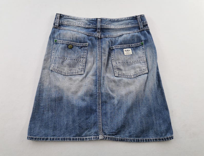 Pu&ograve; includere: Gonna di jeans blu sbiadito con due tasche posteriori e chiusura con bottoni. La gonna ha uno stile classico e casual, con un aspetto leggermente usurato. Le tasche hanno cuciture verdi e una piccola etichetta.