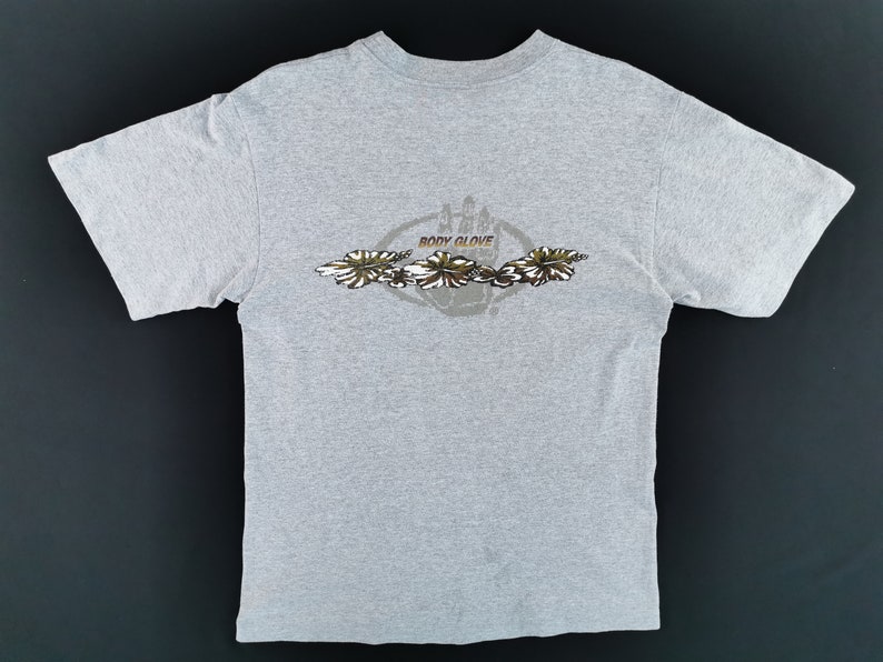 May include: A light gray t-shirt featuring a graphic design on the back. The design includes the words "BODY GLOVE" in a circular emblem, with a floral pattern in shades of brown, white, and green. The shirt is made of a soft material.