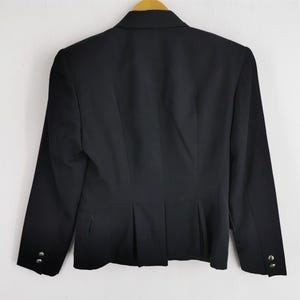 May include: A black blazer with long sleeves, hanging on a wooden hanger. The back of the blazer is visible, showing a tailored fit with a single button closure and decorative buttons on the sleeves. The blazer is made of a smooth, dark fabric.