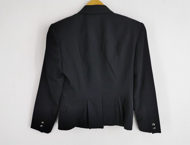 May include: A black blazer with long sleeves, hanging on a wooden hanger. The back of the blazer is visible, showing a tailored fit with a single button closure and decorative buttons on the sleeves. The blazer is made of a smooth, dark fabric.