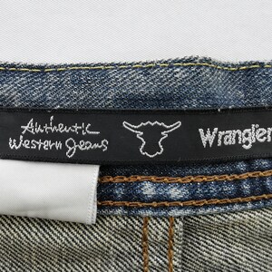 Puede incluir: Primer plano de una etiqueta de vaqueros de mezclilla. La etiqueta negra dice "Authentic Western Jeans" y "Wrangler" en escritura blanca. Se adjunta una etiqueta blanca. Los vaqueros son azules con costuras marrones.