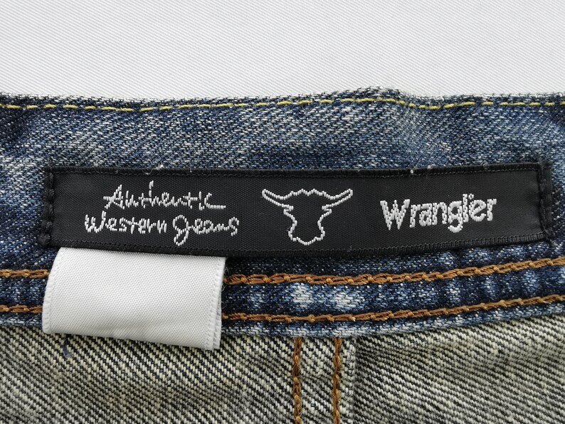 Puede incluir: Primer plano de una etiqueta de vaqueros de mezclilla. La etiqueta negra dice "Authentic Western Jeans" y "Wrangler" en escritura blanca. Se adjunta una etiqueta blanca. Los vaqueros son azules con costuras marrones.
