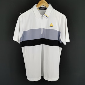 May include: White polo shirt with a color block design featuring a gray and black stripe across the chest. The shirt has a zipper closure and a small yellow logo on the chest. The shirt is hanging on a wooden hanger.