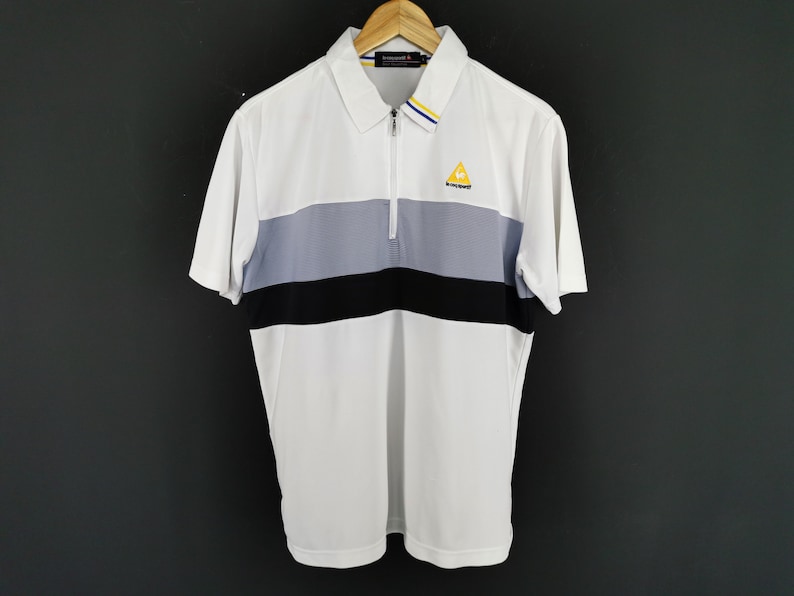 May include: White polo shirt with a color block design featuring a gray and black stripe across the chest. The shirt has a zipper closure and a small yellow logo on the chest. The shirt is hanging on a wooden hanger.