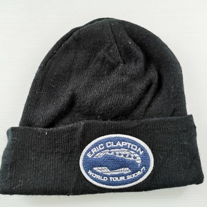 Puede incluir: Gorro de punto negro con borde doblado. Cuenta con un parche con el texto "ERIC CLAPTON WORLD TOUR 2006/7" en letras blancas sobre un fondo azul. El gorro está hecho de una tela suave y texturizada.
