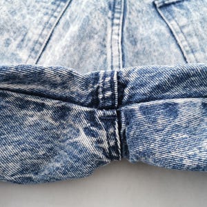 May include: Close-up of acid wash denim jeans. The jeans have a light blue and white pattern, with visible stitching and a small brown patch. The fabric is textured and shows signs of wear.