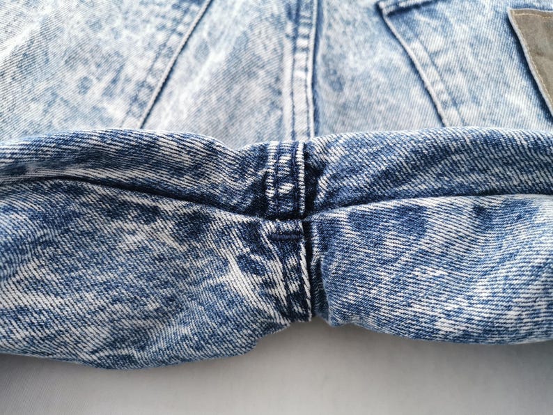 May include: Close-up of acid wash denim jeans. The jeans have a light blue and white pattern, with visible stitching and a small brown patch. The fabric is textured and shows signs of wear.