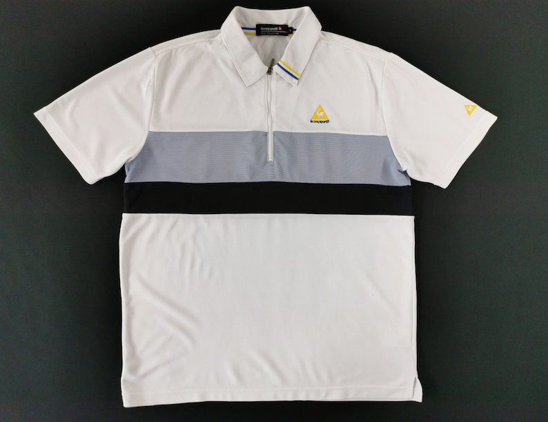 May include: White polo shirt with a color block design featuring a gray and black stripe across the chest. The shirt has a zipper closure, short sleeves, and a small yellow logo on the chest and sleeve.