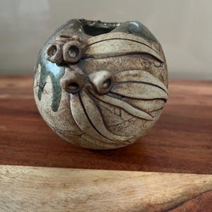 May include: A small, round ceramic vase with a textured, earthy appearance. The vase features raised, leaf-like and bulbous designs in a light beige colour, with dark brown and green accents. The opening is irregular, adding to its unique, handcrafted aesthetic.