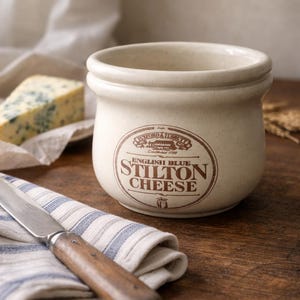 May include: A cream-colored ceramic Stilton cheese crock with a brown logo and text. A blue cheese wedge, a knife, and a striped cloth are on a wooden surface. The crock reads "English Blue Stilton Cheese."