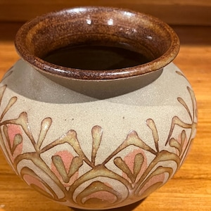 May include: A small, round ceramic vase with a textured, light beige body and a dark brown glazed rim. The exterior features a repeating, hand-painted pattern of brown, leaf-like outlines surrounding soft, muted pink, petal-shaped accents.