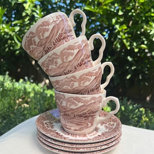 May include: Four brown and white teacups stacked, with matching saucers. Each cup and saucer displays a landscape pattern. The cups have distinctive handles. The set is on a white surface with a green backdrop.