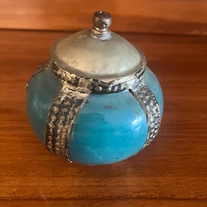May include: A small, round turquoise-colored trinket box with a silver-colored metal band and lid. The lid is a lighter shade of turquoise and has a small knob on top. The box is sitting on a wooden surface.
