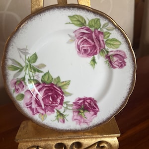 Pode incluir: Um pequeno prato de porcelana branca com borda dourada e um desenho floral de rosas. As rosas são de cor rosa profundo com folhas verdes. O prato é exibido em um pequeno suporte dourado.
