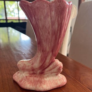 May include: A pink ceramic vase with a unique, trumpet-like shape. The vase has a white base and a pink glaze with a marbled effect.
